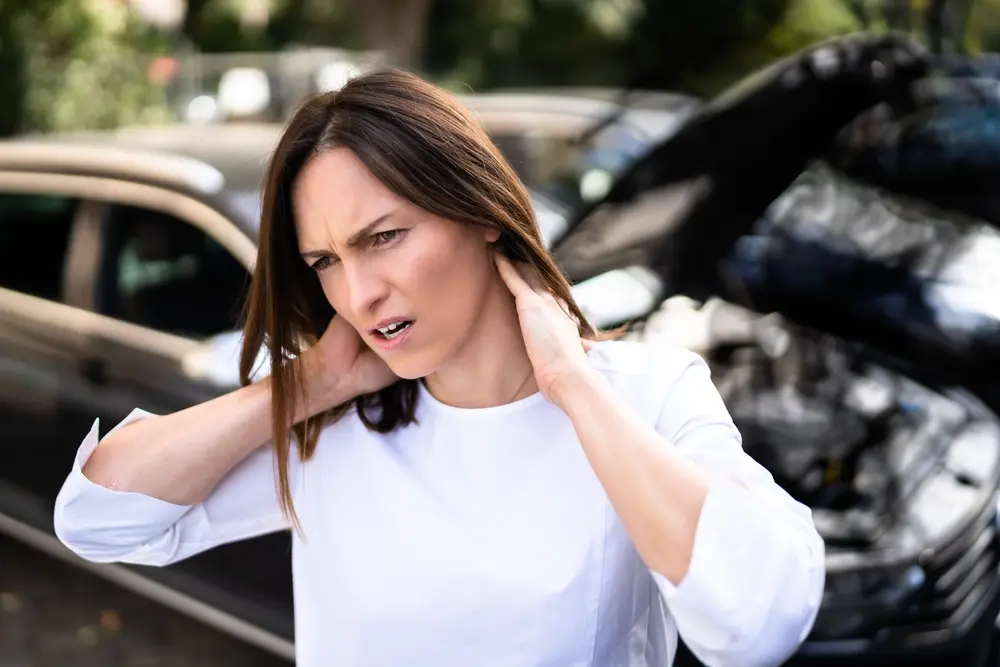 Mujer sujetándose el cuello con dolor tras un accidente de coche, haciendo muecas cerca del vehículo siniestrado con el capó abollado.