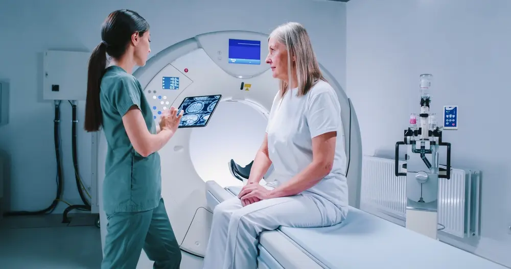 Mujer radióloga explicando los resultados de una resonancia magnética de columna vertebral a una paciente mayor junto a un escáner de resonancia magnética abierta en la sala de diagnóstico.
