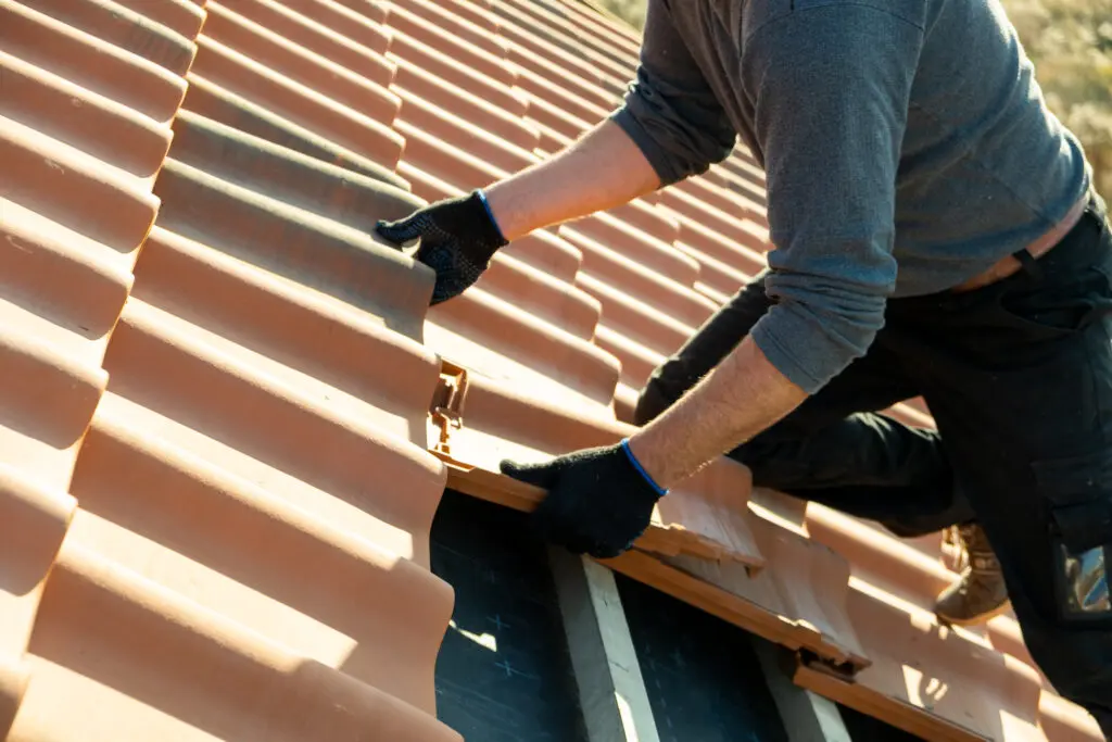 clay tile roof cost Closeup of worker hands installing yellow ceramic roofing tiles mounted on wooden boards covering residential building roof under construction.