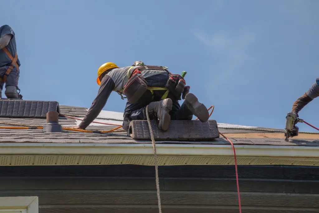 Construction workers wearing safety equipment install asphalt shingles on a residential roof in daylight. Ideal for roofing, home improvement, safety training, and construction industry visuals.