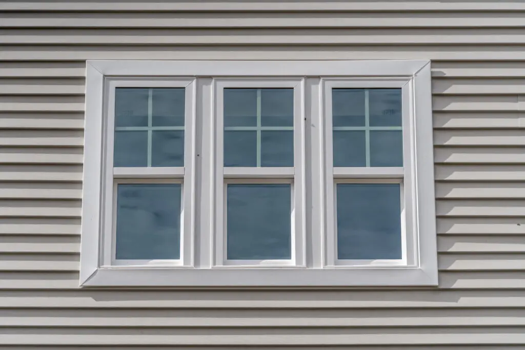 Triple hung window with fixed top sash and bottom sash that slides up, sash divided by white grilles a surrounded by white elegant frame horizontal white vinyl siding on a new construction residence