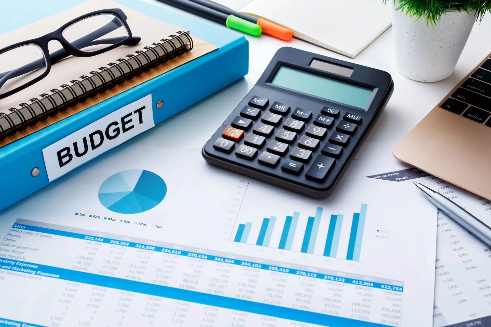 A blue binder labeled Budget sits beside a calculator, eyeglasses and printed charts on an office desk.