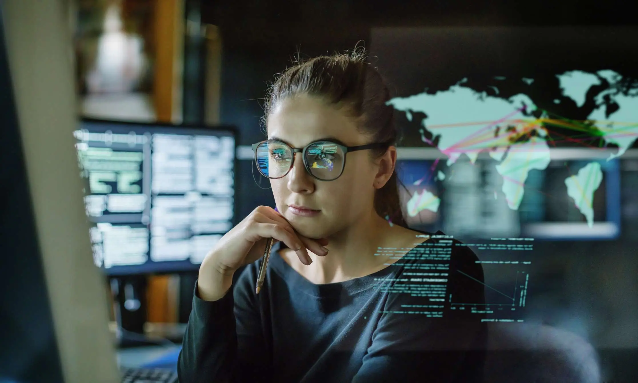 A woman in glasses studies computer screens showing code and a world map in a dim office.
