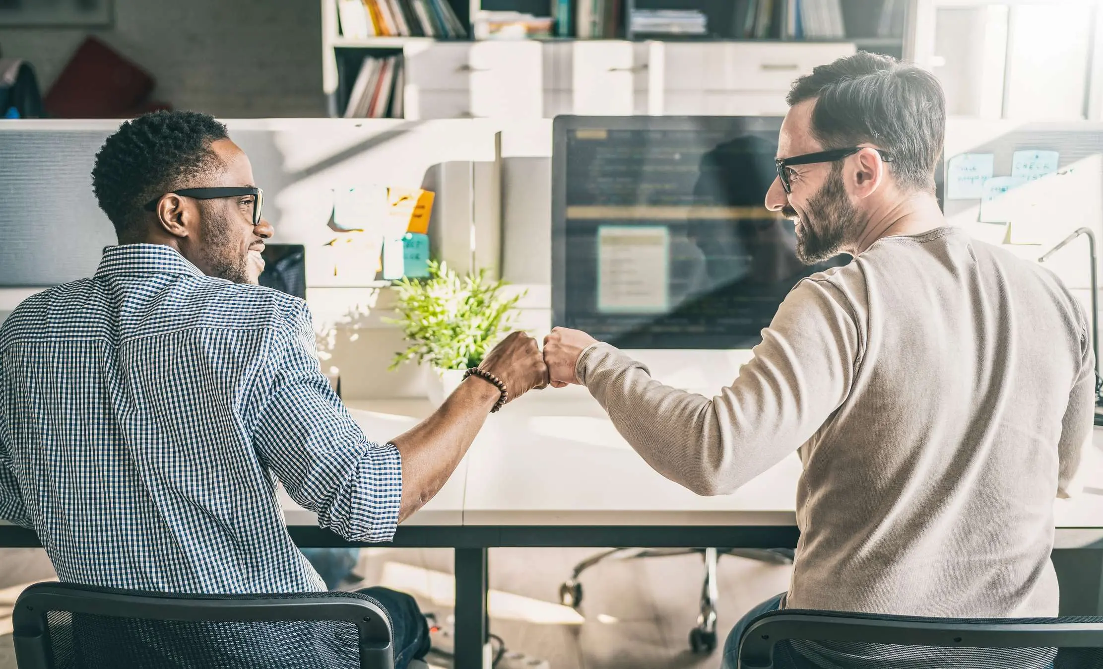 Two coworkers bump fists across a desk in a bright, modern office.
