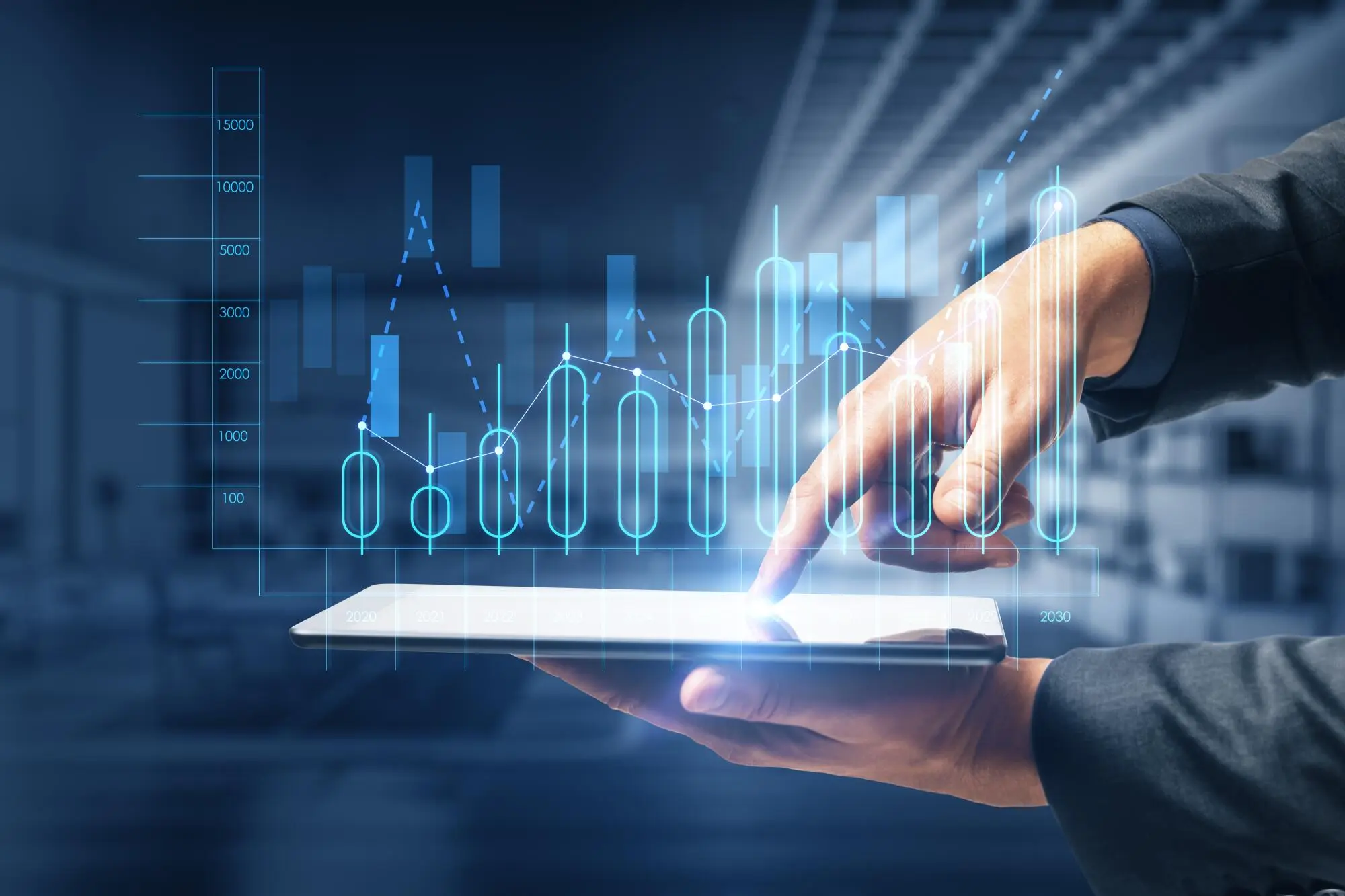 A person in a dark suit holds a white tablet while pointing at blue digital charts and graphs floating above it.