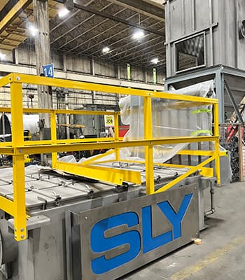 Industrial factory floor with a gray machine surrounded by bright yellow safety railing and a large blue SLY logo on the side.