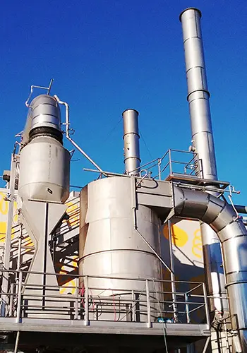 food-manufacturing-emissions | Sly Air Pollution Control Industrial processing plant with tall silver chimneys, large cylindrical tanks, and a network of pipes on a raised platform against a blue sky.