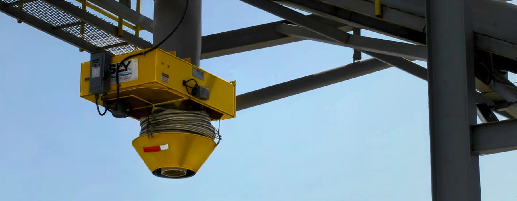 Durable-Loading-Spouts-Hero | Sly Air Pollution Control Yellow industrial hoist with a spool of cable attached to a steel framework against a blue sky, likely part of an overhead crane system.