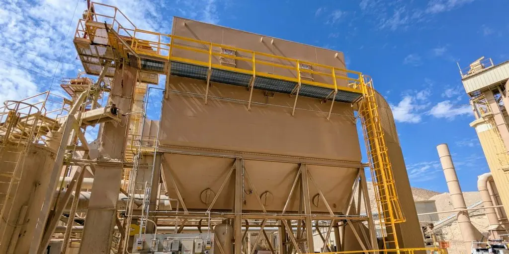 Industrial processing plant with beige/bronze metal structures, yellow safety rails, and a platformed conveyor under a blue sky.