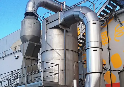 food-manufacturing-scrubber-emissions | Sly Air Pollution Control Industrial piping and metal ducts on an outdoor factory facade with stairs and platforms nearby, against a blue sky.