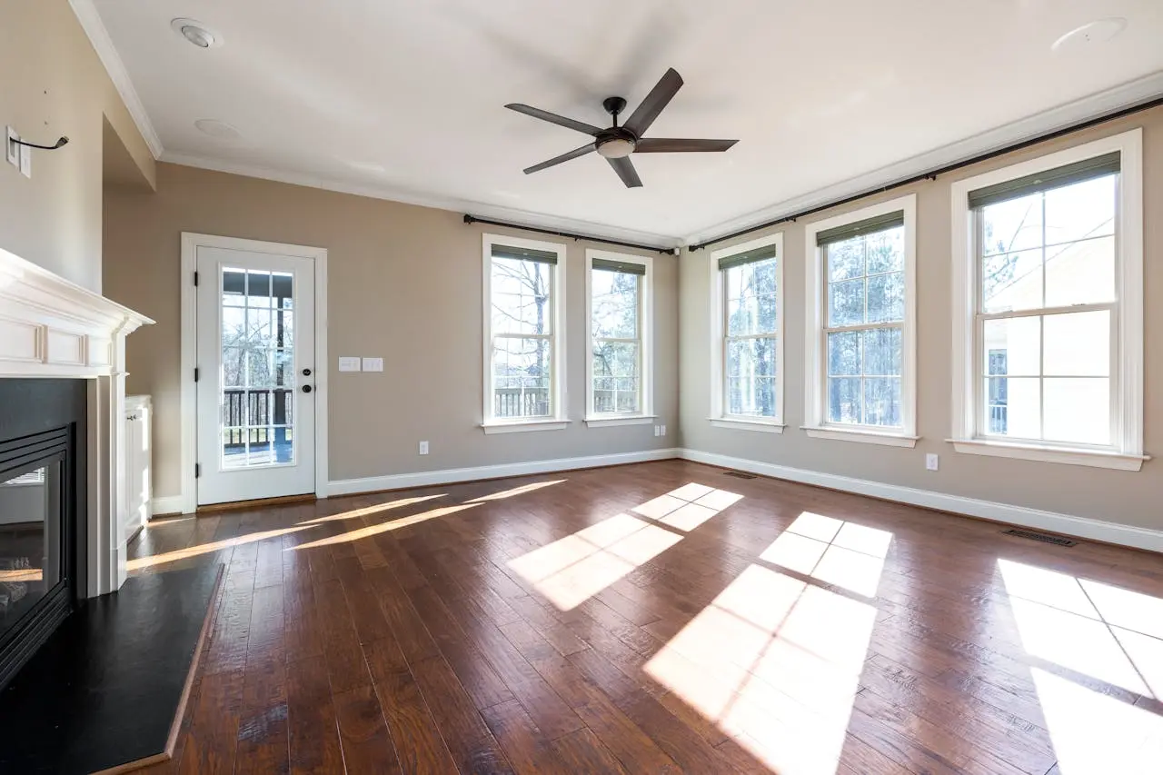 Bright empty room with hardwood floors, large windows, ceiling fan, and a fireplace.