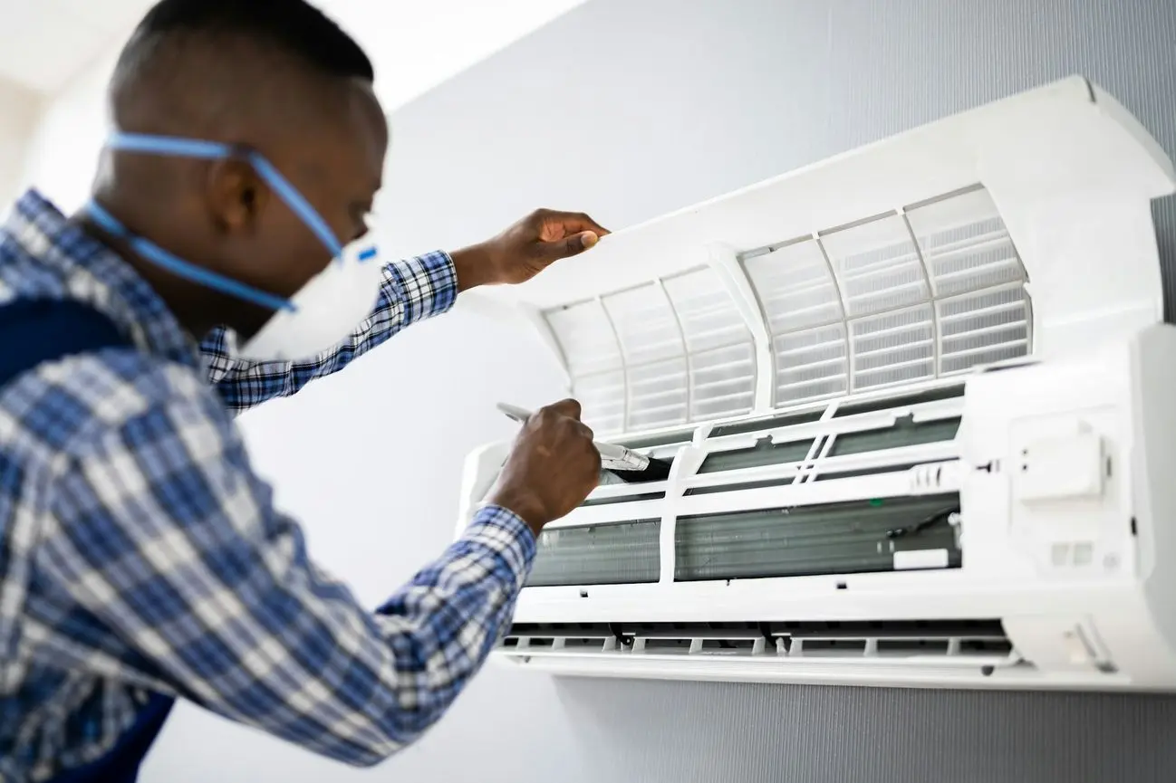 Technician wearing a mask cleans the interior of a wall-mounted air conditioner with a brush.