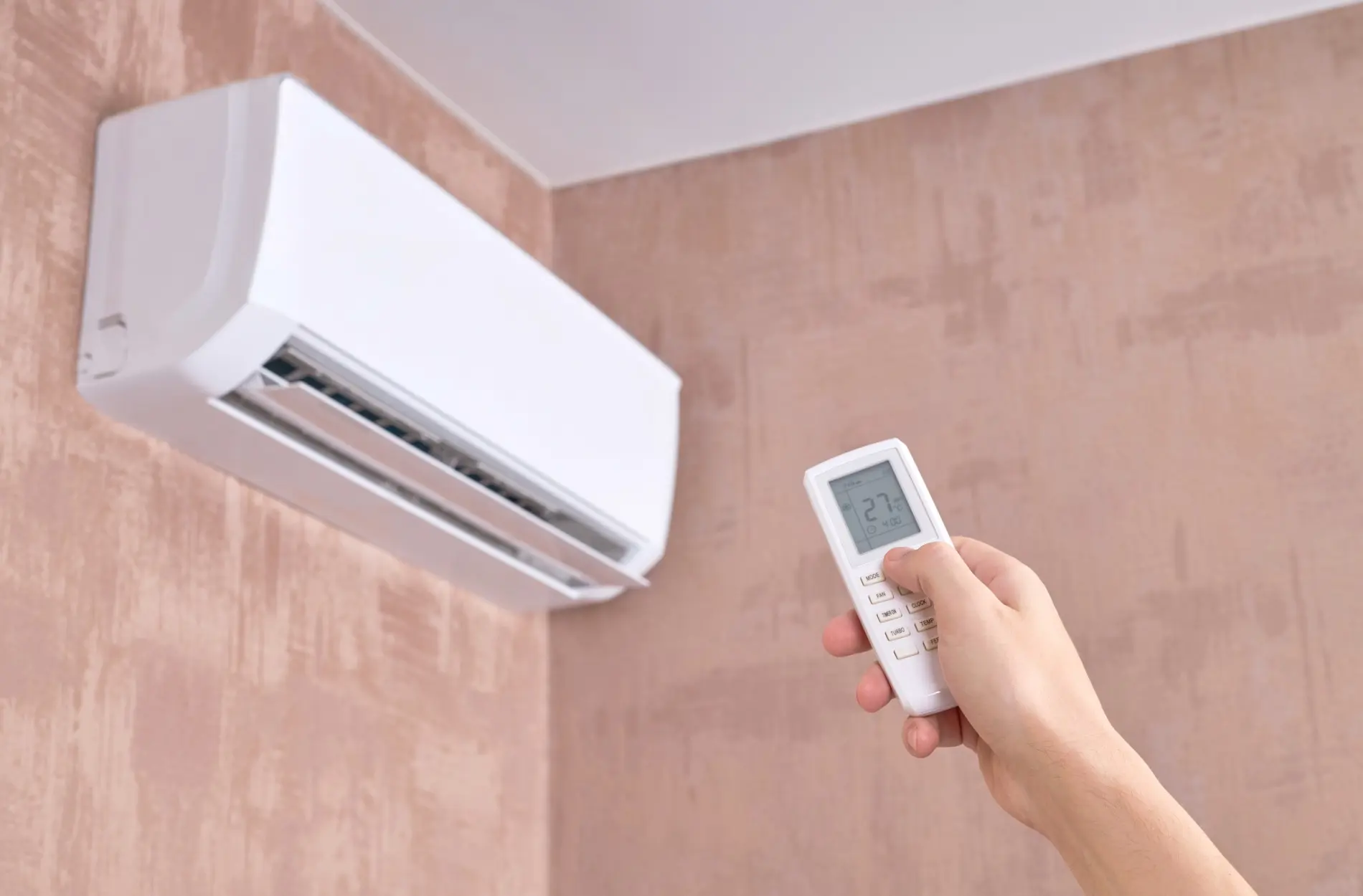 Hand holding remote control towards wall-mounted air conditioning unit.