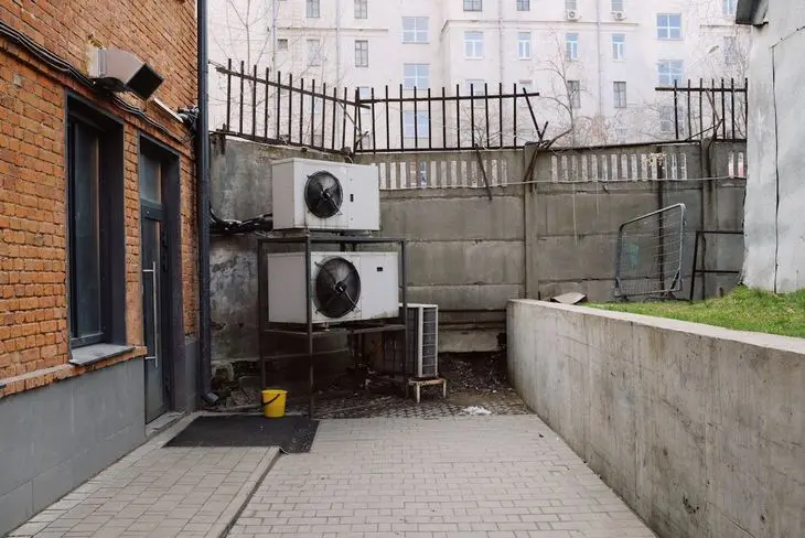 Outdoor air conditioning units against a brick building wall, with a metal fence in the background.