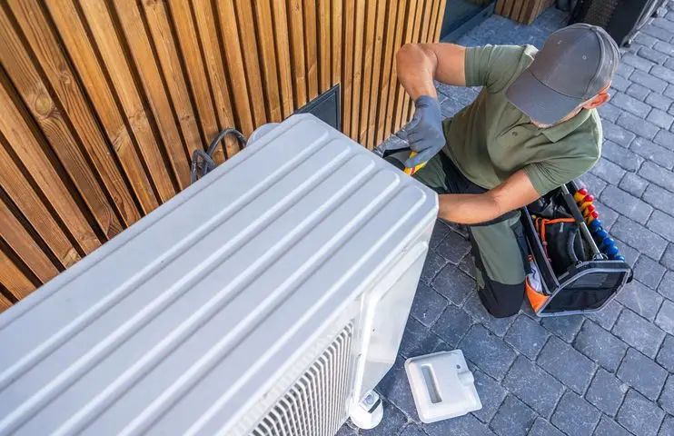 Technician in a cap and gloves repairs an outdoor air conditioning unit, with tools and a toolbox nearby on the ground.