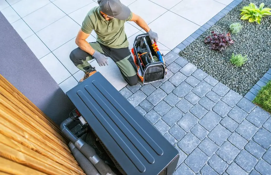 Technician kneeling beside an HVAC unit with a toolkit on a paved patio.