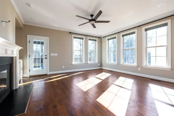 Bright empty room with hardwood floors, large windows, ceiling fan, and a fireplace.
