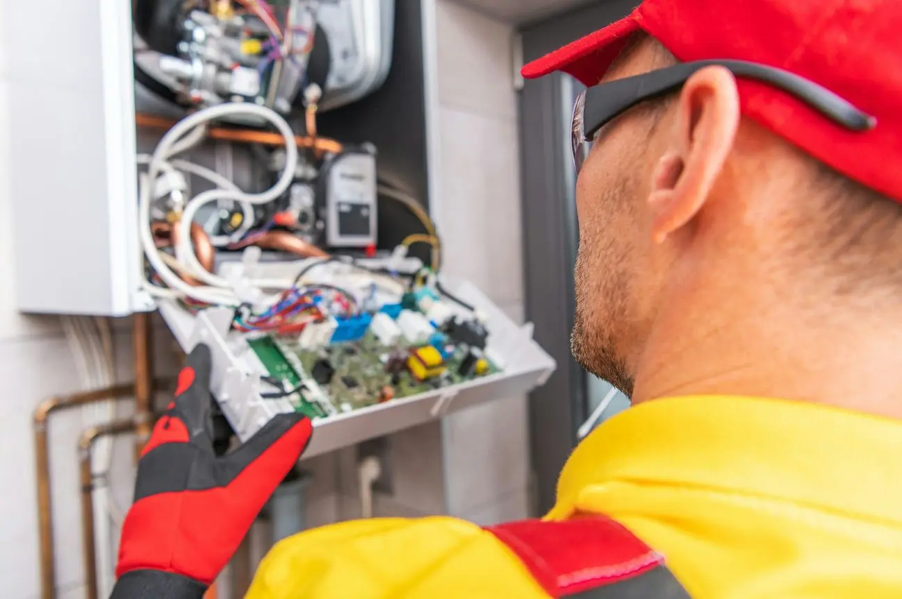 Technician in red cap and gloves inspecting HVAC system with exposed wires and components.