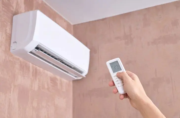 Hand holding remote control towards wall-mounted air conditioning unit.