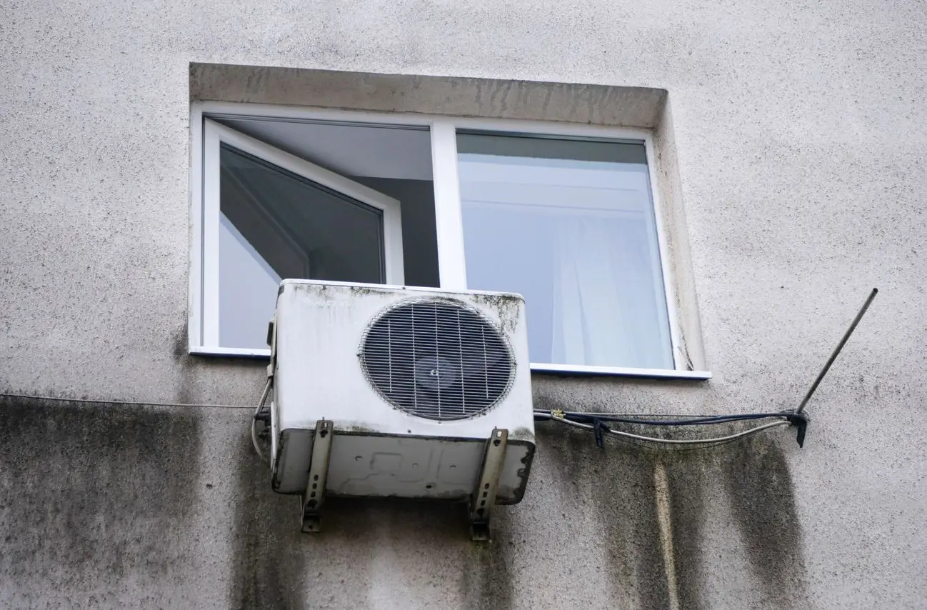 Wall-mounted air conditioning unit below an open window on a concrete building facade.