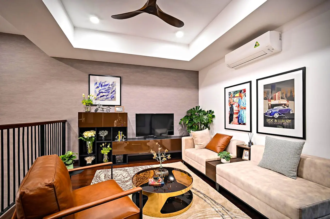Modern living room with a beige sofa, brown armchair, wall art, TV, plants, and a glass coffee table beneath a ceiling fan.