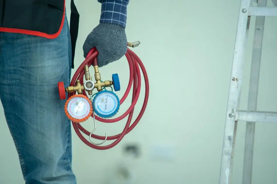 Technician holding HVAC manifold gauge set with red hoses next to a metal ladder.