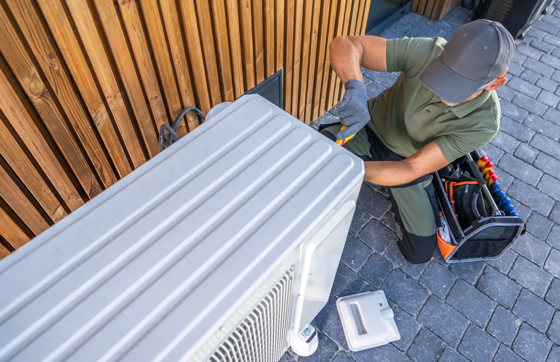Technician in a cap and gloves repairs an outdoor air conditioning unit, with tools and a toolbox nearby on the ground.