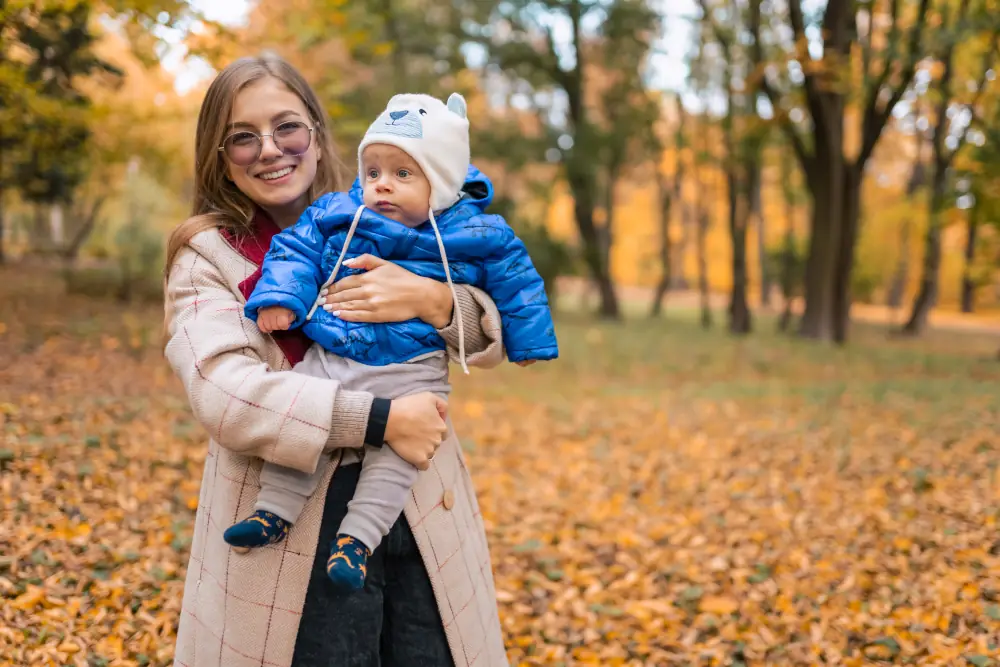 A smiling woman holding a baby in a blue jacket and hat, standing in a park with fallen autumn leaves and trees in the background.
