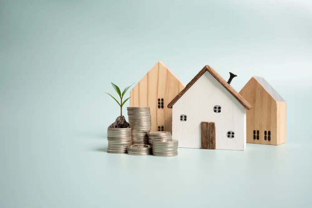 Wooden house models beside stacked coins and a small plant sprouting from a coin pile.