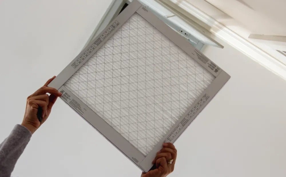 Hands holding an air filter near a ceiling vent.