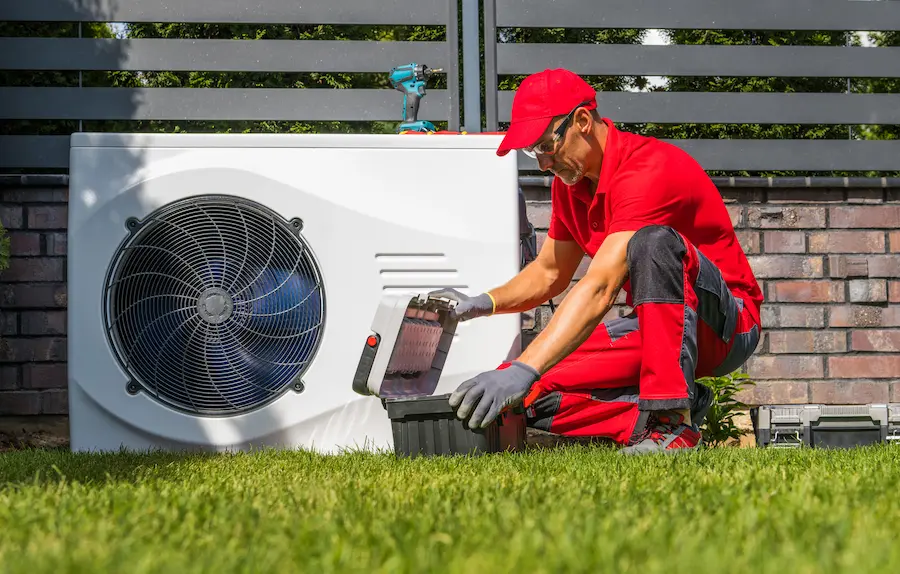 Technician in red uniform working on an outdoor HVAC unit, opening a black toolbox on a grassy lawn.