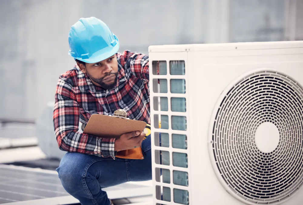 Technician in a blue hard hat inspecting an outdoor air conditioning unit while holding a clipboard.