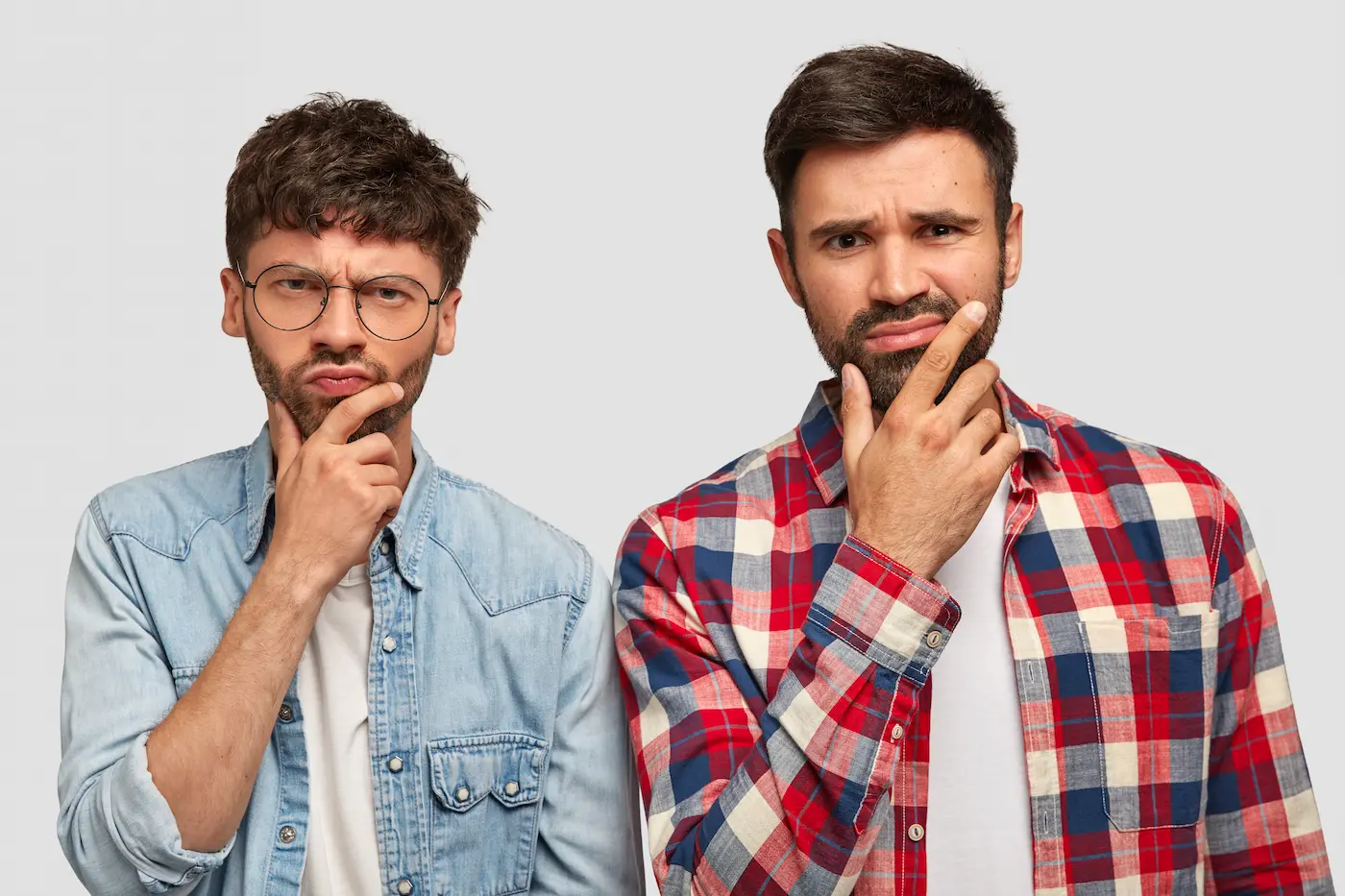 Two men in casual shirts express thoughtful, puzzled expressions while touching their chins against a plain background.