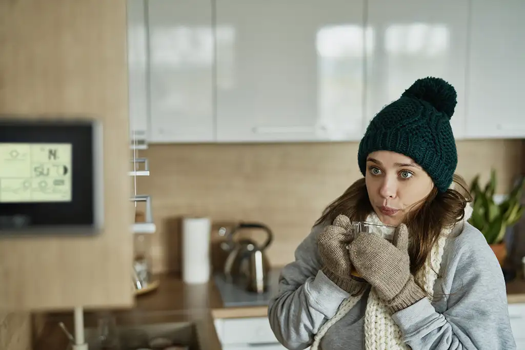 Person in a green beanie and gloves drinking tea in a cozy kitchen with a thermostat on the wall.