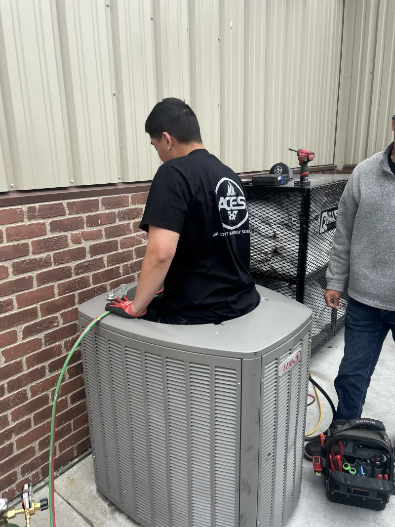 Technician servicing an outdoor AC unit beside a brick wall, with tools and a toolkit nearby.