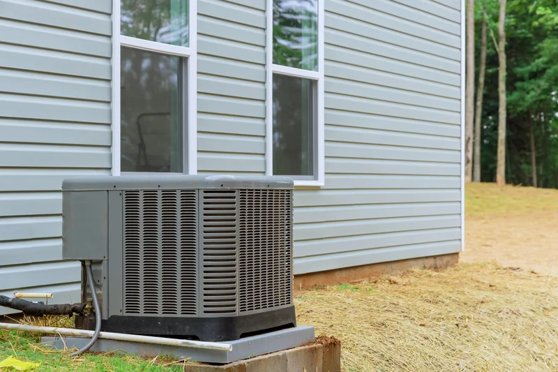Outdoor central air conditioning unit next to a house with gray siding and two windows.