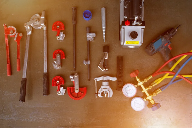 Assorted HVAC tools including pipe benders, cutting tools, a drill, and pressure gauges arranged on a dark surface.