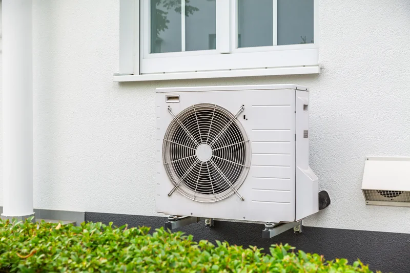 Outdoor air conditioning unit mounted on a white wall beneath a window, surrounded by green shrubs.