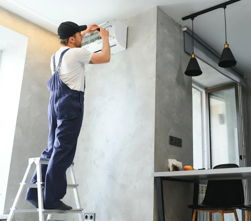 Technician in overalls on a ladder fixing a wall-mounted air conditioning unit in a modern room.