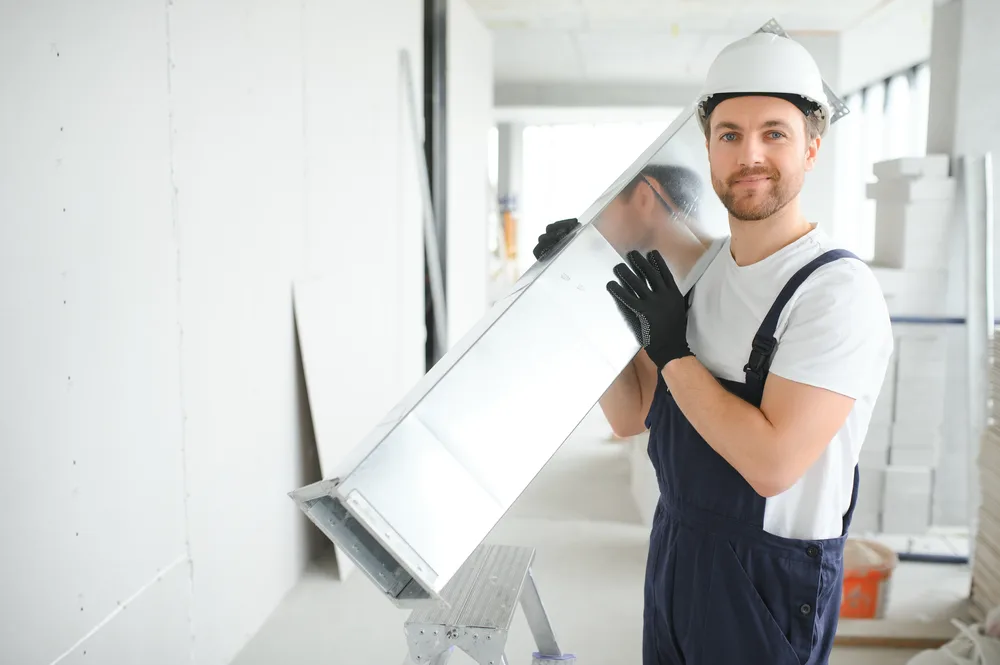 Worker in a hard hat holding a metal duct in a bright, unfinished room.
