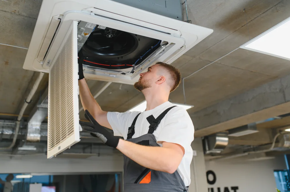 Technician adjusting a ceiling-mounted HVAC unit in an industrial setting.