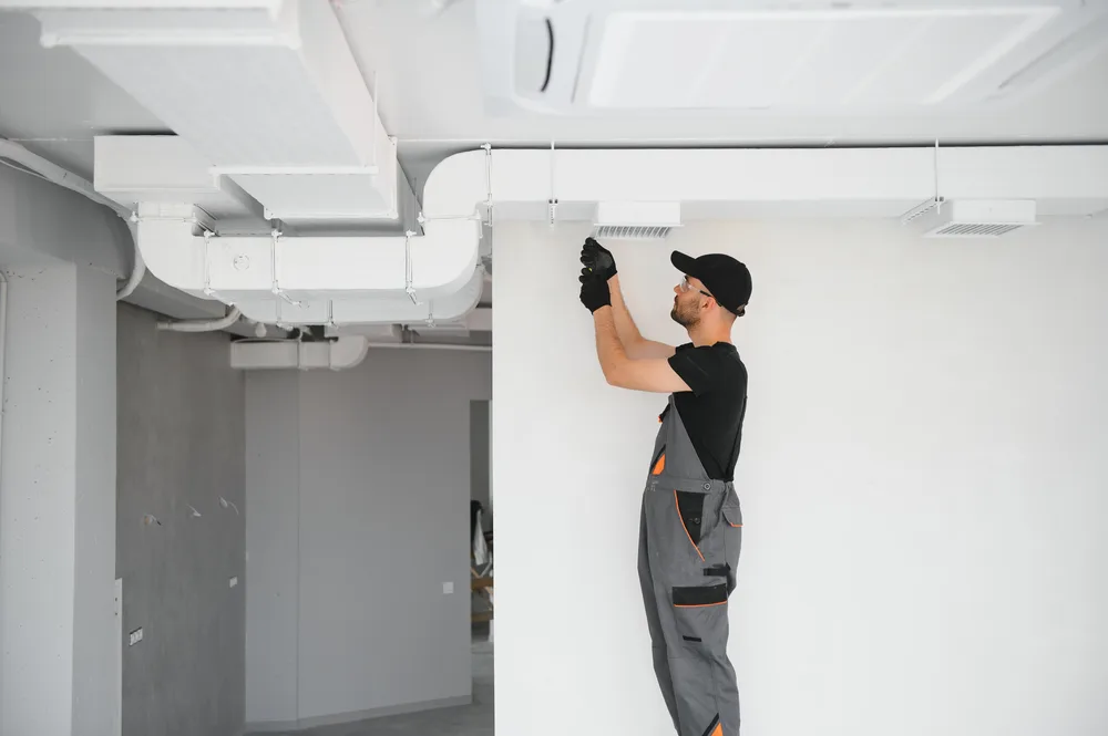 Technician in overalls and cap installing or maintaining an HVAC system on a white ceiling.