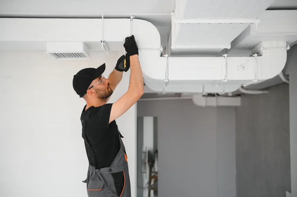Technician adjusting ductwork on a ceiling wearing black cap and gloves.