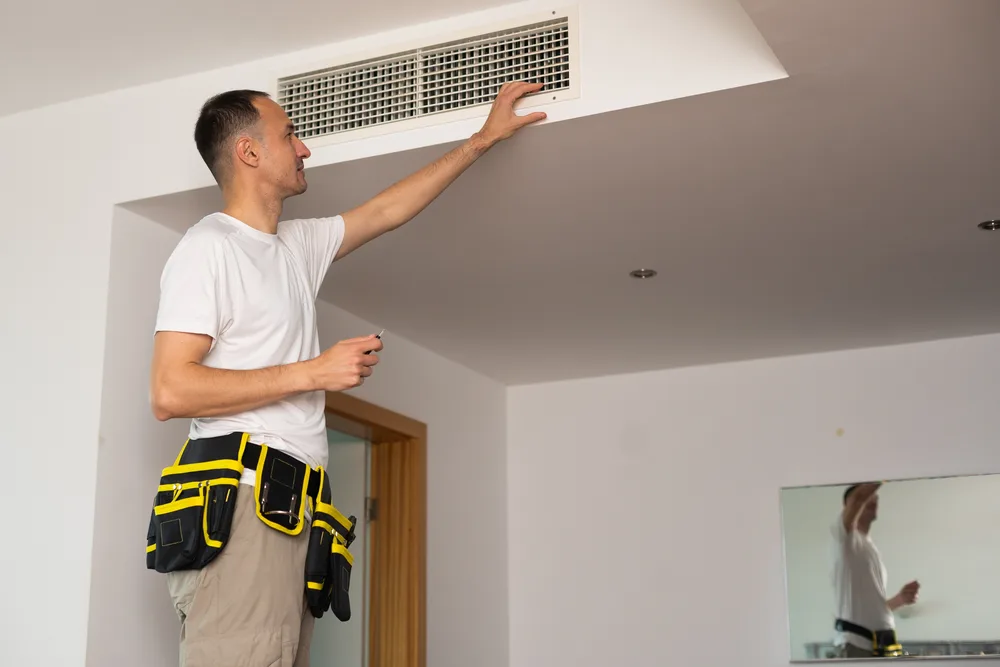 Technician inspecting ceiling air vent while holding a tool, wearing a tool belt.