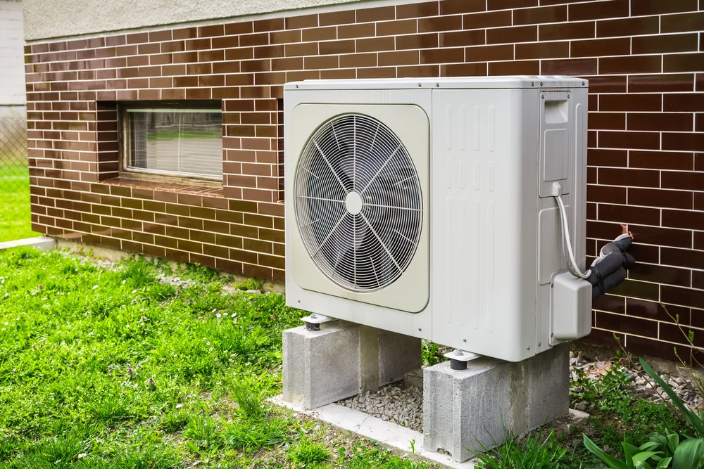 Outdoor air conditioning unit mounted on concrete blocks beside a brick wall with a window.