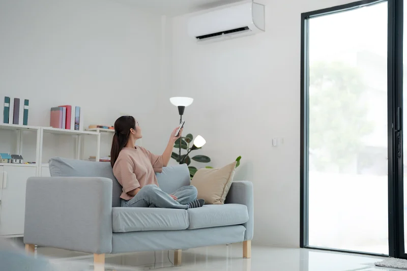 Woman sitting on a couch using a remote to adjust the air conditioner in a bright, modern living room.