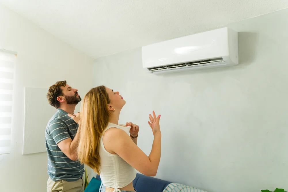 Two people standing in a room enjoying cool air from a wall-mounted air conditioning unit.