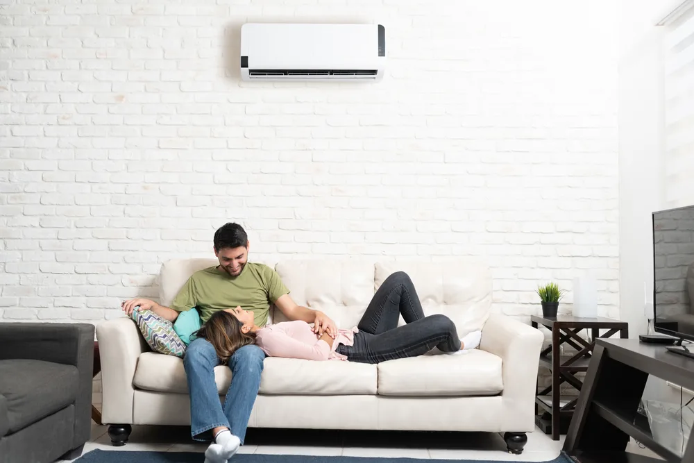 Couple relaxing on a beige sofa under a wall-mounted air conditioner in a cozy living room.