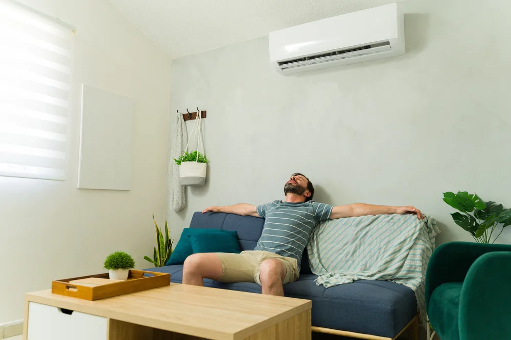Man relaxing on a blue couch under a running air conditioner in a cozy living room.