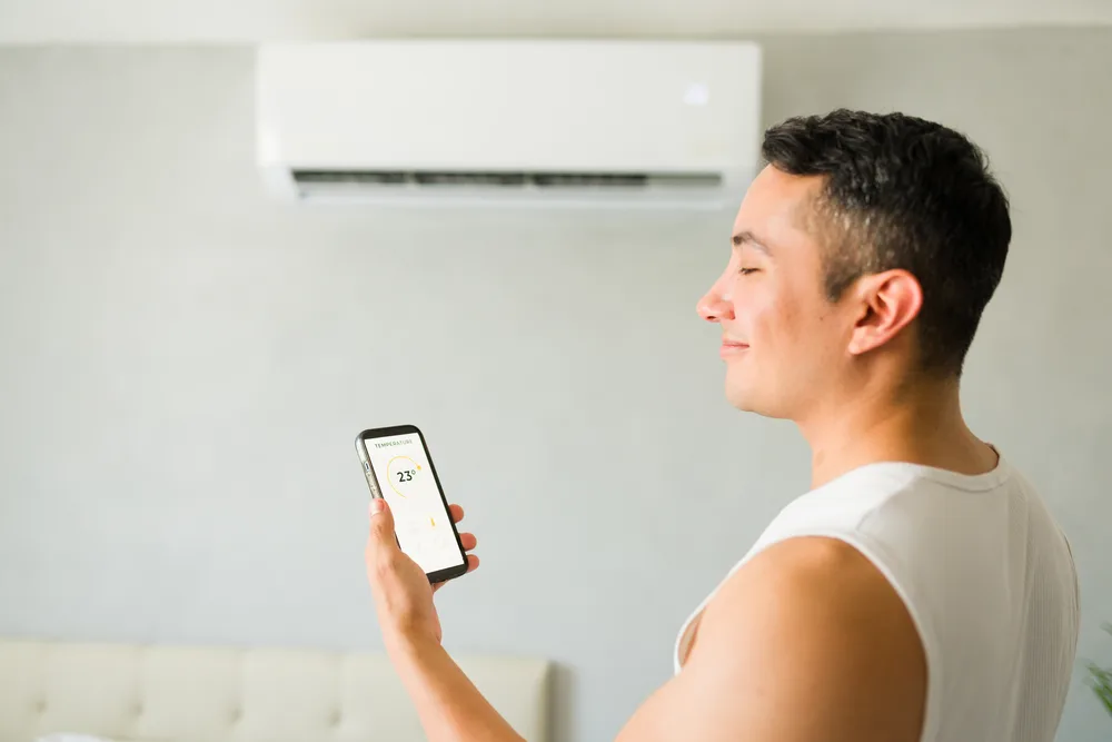 Man adjusting air conditioner with a smartphone, displaying a temperature of 23 degrees.